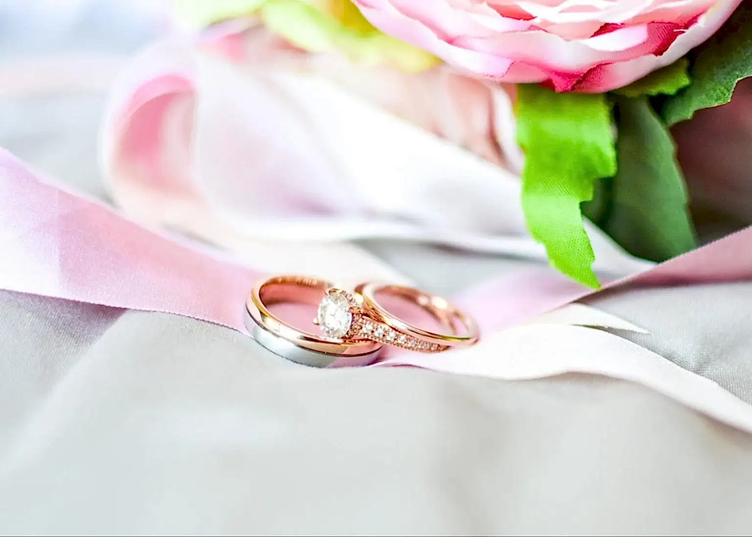 A pair of channel-set engagement rings sit on a pink bow atop a white tablecloth with a pink rose visible behind.