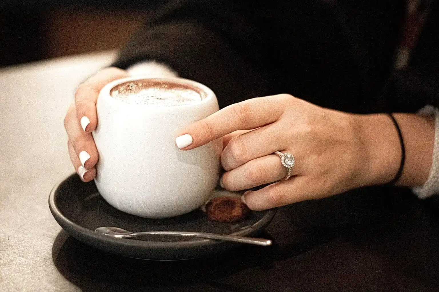 A woman with white fingernails sips coffee from a small white mug, with the halo ring on her ring finger clearly visible.