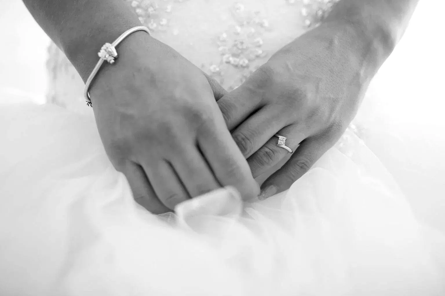 A black and white photo showing a bride's hands linked in front of her dress, with a tension engagement ring visible on her ring finger.