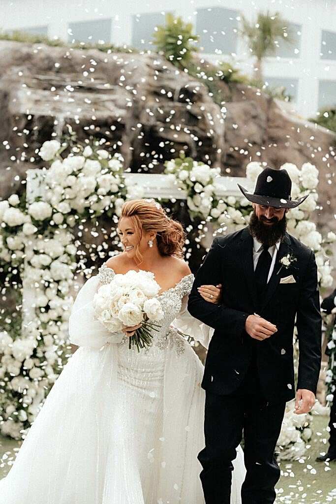 A bride holds a man's arm as she walks down the aisle and white flower petals are thrown around her
