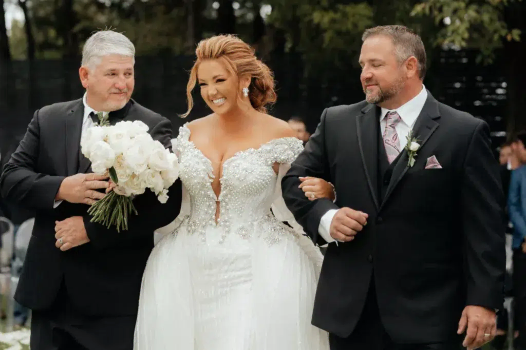 A bride is accompanied by her father and the groom as they walk the aisle. She's wearing a bespoke tailored wedding dress created just for her.