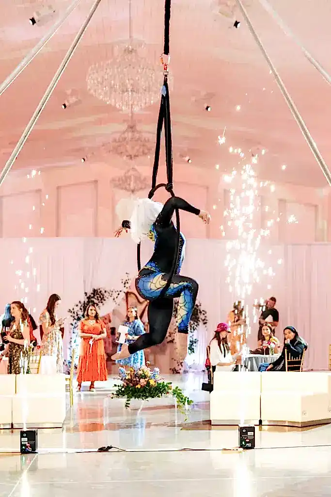 An acrobat performs a routine using hanging silk ropes in a wedding venue with pyrotechnics and guests in the background.