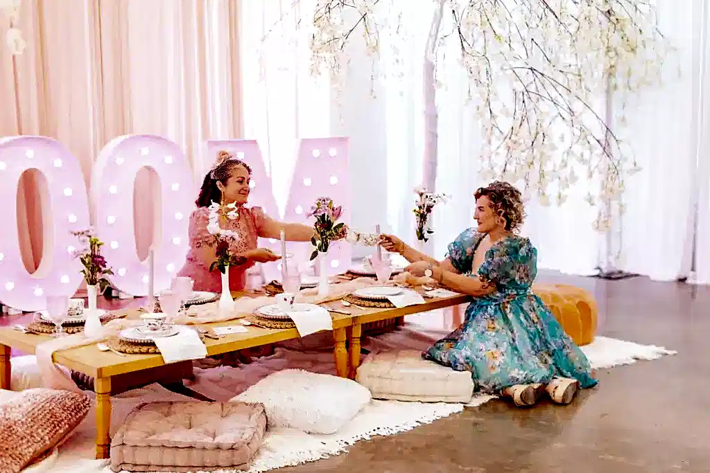 Guests at a wedding sit next to a wall that uses a tree for decoration. The tree is wrapped in light pink string with lightly colored leaves and small pink-white flower blossoms.